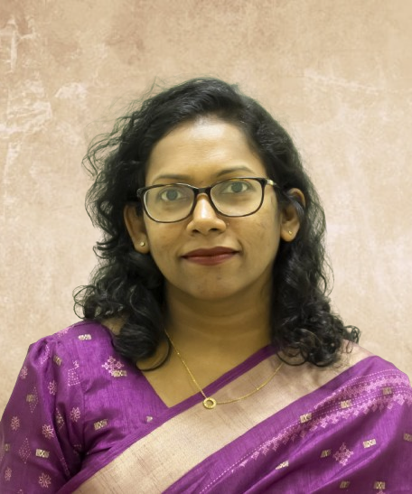 Portrait of a woman with glasses wearing a purple saree and gold necklace, facing the camera.
