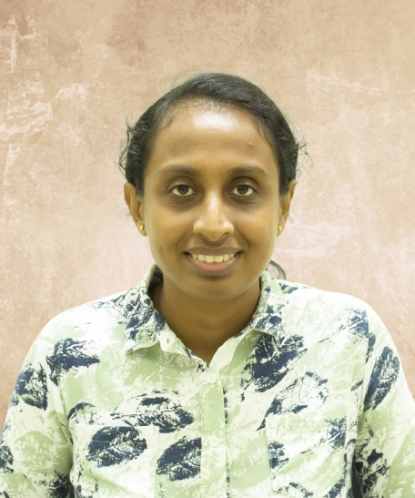Portrait of a smiling woman with dark hair in a light green patterned shirt, standing against a beige wall.