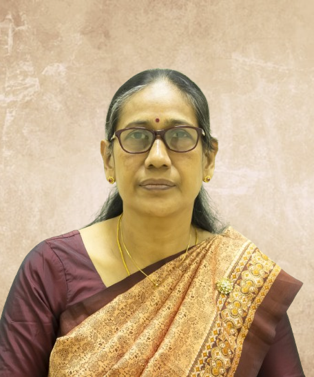 Portrait of an Indian woman facing the camera, wearing a maroon blouse and gold silk saree with a bindi and glasses, gold earrings, and a necklace.