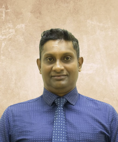 Portrait of a man in a blue shirt and patterned tie, standing against a beige textured wall.