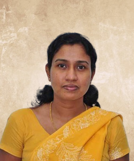 Portrait of a woman in a yellow sari with a gold necklace against a beige textured wall, facing the camera.